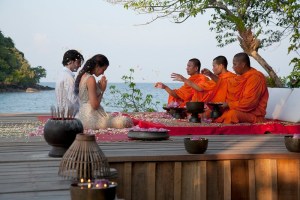 Man and woman being blessed by three monks in orange robes by the ocean in Cambodia